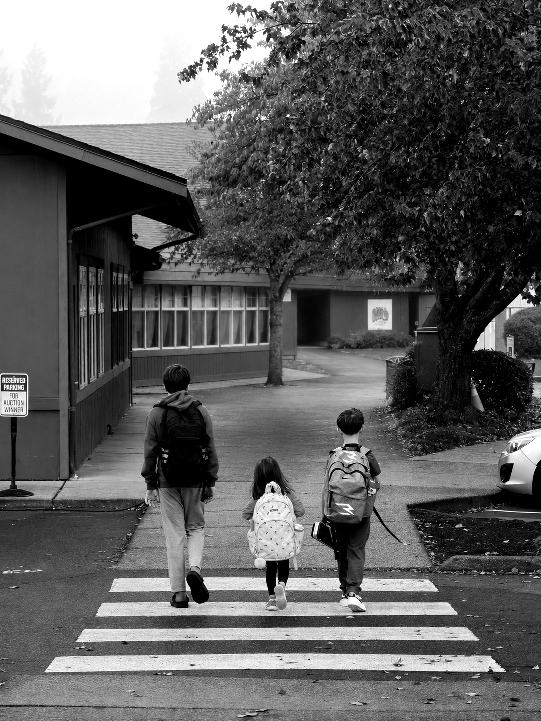 Three people, two children and an adult with school backpacks, cross a road on a marked zebra crossing, seen from a distance.
