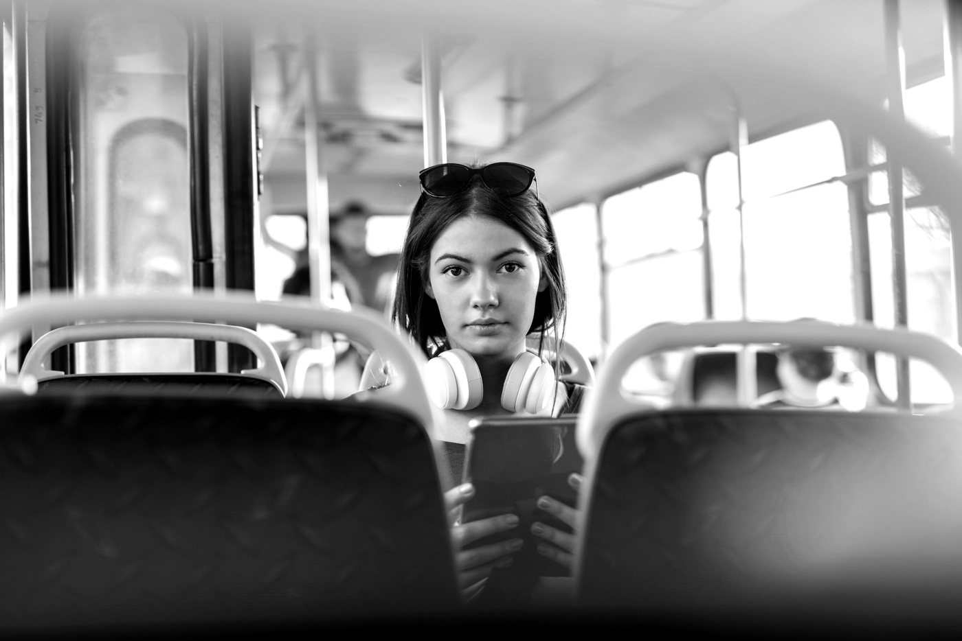 A young woman sits on a bus, headphones round her neck, looking directly at the camera.